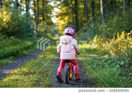 Child riding balance bike. Kids on bicycle in sunny forest. Little girl enjoying to ride glider bike on warm day. Preschooler learning to balance on run bicycle in safe helmet. Sport activity Child riding balance bike. Kids on bicycle in sunny forest. Little girl enjoying to ride glider bike on warm day. Preschooler learning to balance on run bicycle in safe helmet. Sport activity 108680680