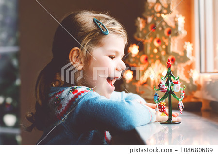 Litte toddler girl sitting by window and decorating small glass Christmas tree with tiny xmas toys. Happy healthy child celebrate family traditional holiday. Adorable baby. Litte toddler girl sitting by window and decorating small glass Christmas tree with tiny xmas toys. Happy healthy child celebrate family traditional holiday. Adorable baby. 108680689
