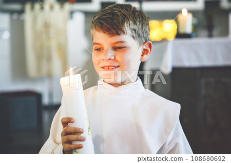 Little kid boy receiving his first holy communion. Happy child holding Christening candle. Tradition in catholic curch. Kid in a white traditional gown in a church near altar. Little kid boy receiving his first holy communion. Happy child holding Christening candle. Tradition in catholic curch. Kid in a white traditional gown in a church near altar. 108680692