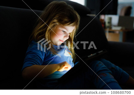 Cute little toddler girl playing with tablet pc at home. Healthy baby touching pad with fingers, looking cartoons and having fun with educational apps. Child in pajamas nightwear hear audio book 108680700