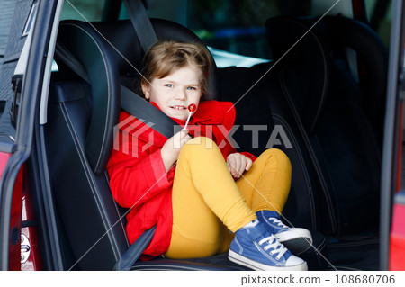 Little toddler girl sitting in car seat, holding sweet lollipop against nausea and sickness and looking out of the window on nature and traffic. Cute kid traveling by car. Child safety on the road. Little toddler girl sitting in car seat, holding sweet lollipop against nausea and sickness and looking out of the window on nature and traffic. Cute kid traveling by car. Child safety on the road. 108680706