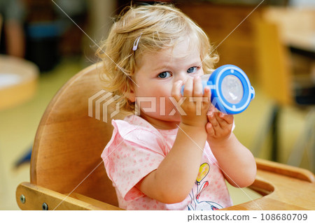 Adorable toddler girl drinking formula milk or water from bottle. Cute happy baby child taking food from dish at daycare or nursery canteen. 108680709