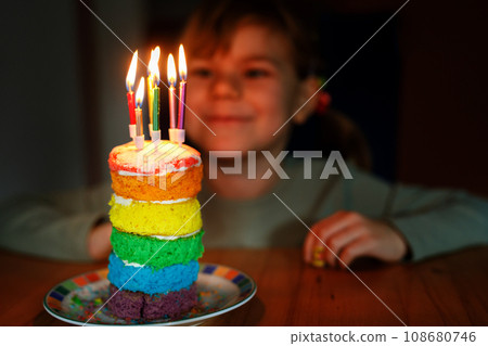 Happy little preschool girl celebrating birthday. Closeup of child with homemade rainbow cake, indoor. Happy healthy toddler blowing six candles on cake. Selective focus on cake Happy little preschool girl celebrating birthday. Closeup of child with homemade rainbow cake, indoor. Happy healthy toddler blowing six candles on cake. Selective focus on cake 108680746