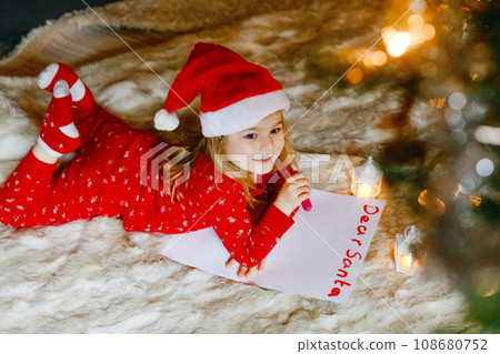 Little cute preschool girl in pajamas under Christmas tree, writing wish list letter to Santa Claus at home, indoors. Traditional Christian festival. Happy toddler child waiting for gifts on xmas. 108680752