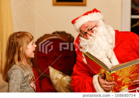 Little toddler girl talking to Santa Claus called Nikolaus or Weihnachtsmann in German. Happy smiling kid waiting for gifts. Santa with golden book. Xmas, childhood, holiday Little toddler girl talking to Santa Claus called Nikolaus or Weihnachtsmann in German. Happy smiling kid waiting for gifts. Santa with golden book. Xmas, childhood, holiday 108680754