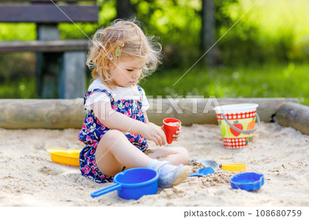 Cute toddler girl playing in sand on outdoor playground. Beautiful baby in red trousers having fun on sunny warm summer day. Child with colorful sand toys. Healthy active baby outdoors plays games Cute toddler girl playing in sand on outdoor playground. Beautiful baby in red trousers having fun on sunny warm summer day. Child with colorful sand toys. Healthy active baby outdoors plays games 108680759
