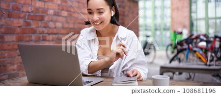 Smiling asian girl in wireless earphones, looking at laptop screen, video chat, working online from coworking, sitting in office Smiling asian girl in wireless earphones, looking at laptop screen, video chat, working online from coworking, sitting in office 108681196