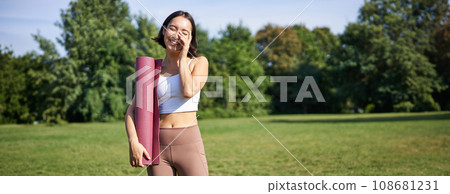 Portrait of asian girl smiles and laughs, stands with sports equipment, rubber mat, wears uniform for workout and exercises in park 108681231