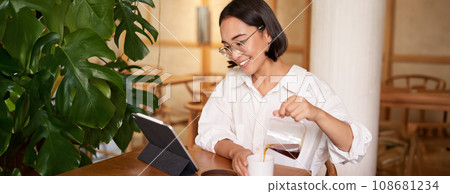 Freelance and remote workers. Smiling young woman pouring coffee in a cup, sitting in cafe and looking at digital tablet Freelance and remote workers. Smiling young woman pouring coffee in a cup, sitting in cafe and looking at digital tablet 108681234