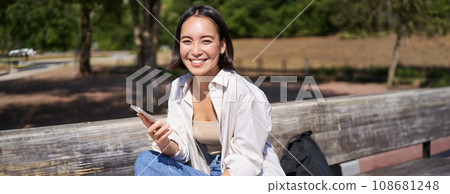 Portrait of happy young asian woman talking on mobile phone in park, sitting on bench and having a telephone call, chatting lively Portrait of happy young asian woman talking on mobile phone in park, sitting on bench and having a telephone call, chatting lively 108681248