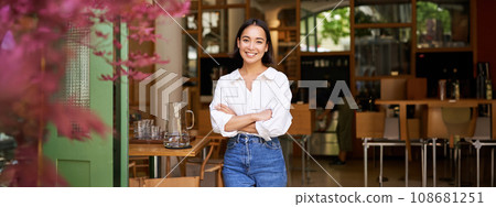 Portrait of young businesswoman in her own cafe, manager standing near entrance and inviting you, posing in white plain shirt and jeans 108681251
