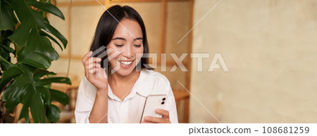Portrait of modern girl sits with coffee in cafe, smiling while looking at smartphone, reading book in restaurant and using mobile phone app Portrait of modern girl sits with coffee in cafe, smiling while looking at smartphone, reading book in restaurant and using mobile phone app 108681259