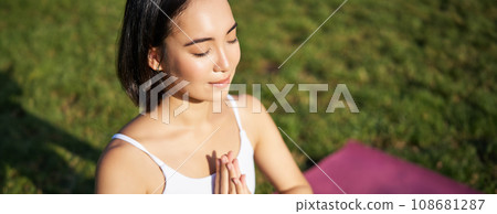 Portrait of young mindful woman, practice yoga, exercising, inhale and exhale on fresh air in park, sitting on rubber mat 108681287