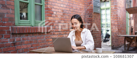 Happy asian girl watching video on laptop, doing homework outdoor in coworking space, smiling and working, drinking coffee 108681307