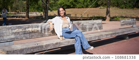 Portrait of beautiful young woman sitting on bench in park, using laptop and drinking coffee, relaxing outdoors on summer day 108681338
