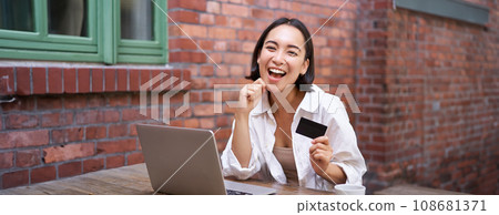 Happy young asian woman sitting near laptop, holding credit card, paying bills, shopping online contactless, smiling at camera 108681371