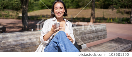 Happy smiling asian girl sitting on bench in park and browsing on social media, holding smartphone, using mobile phone app 108681390