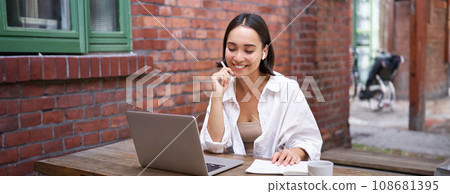 Young smiling asian woman with wireless earphones, sitting in coworking office space with laptop, doing homework, making notes 108681395