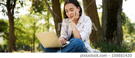 Portrait of asian woman sitting on grass near tree, using laptop, working, doing homework remotely on sunny summer day Portrait of asian woman sitting on grass near tree, using laptop, working, doing homework remotely on sunny summer day 108681420