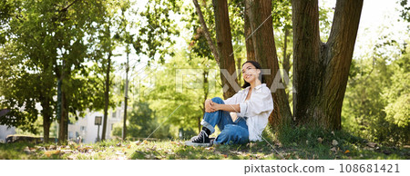 Beautiful smiling girl sits near tree in park, enjoying nature outdoors, relaxing and resting on fresh air Beautiful smiling girl sits near tree in park, enjoying nature outdoors, relaxing and resting on fresh air 108681421