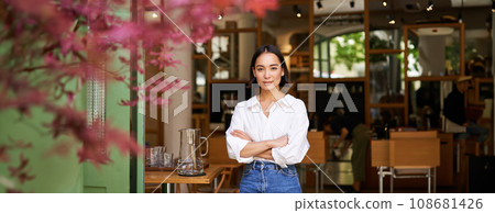 Portrait of young businesswoman in her own cafe, manager standing near entrance and inviting you, posing in white plain shirt and jeans 108681426