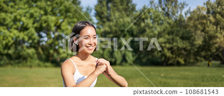 Vertical shot of young fit woman does squats in park, using stretching band on legs, smiling pleased while workout 108681543