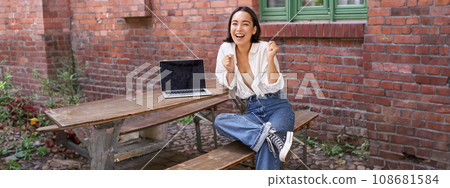 Vertical shot of enthusiastic asian girl, sits with laptop, screen is empty and blank, woman laughing and celebrating, triumphing 108681584