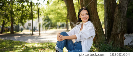 Beautiful smiling girl sits near tree in park, enjoying nature outdoors, relaxing and resting on fresh air 108681604