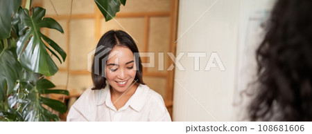 Happy asian woman sits in cafe with cup of coffee and book, answer video call on smartphone, laughing and talking via camera on mobile phone 108681606