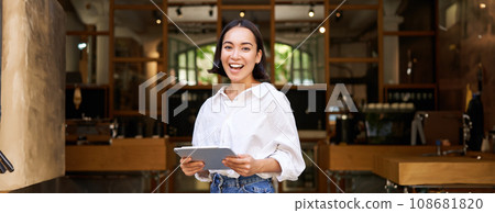 Smiling beautiful waitress, businesswoman with digital tablet, laughing and looking happy, standing in front of cafe or restaurant Smiling beautiful waitress, businesswoman with digital tablet, laughing and looking happy, standing in front of cafe or restaurant 108681820