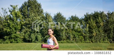 Vertical shot of stylish fitness woman with rubber mat, posing on lawn in park, doing workout on fresh air Vertical shot of stylish fitness woman with rubber mat, posing on lawn in park, doing workout on fresh air 108681832