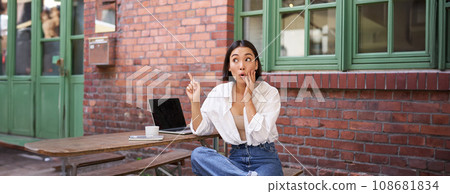 Stylish urban girl, asian woman with laptop sits in cafe, points at upper right corner banner, shows copy space with surprised face expression 108681834