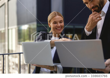 Smiling business colleagues discuss biz issue while use laptop standing on office terrace Smiling business colleagues discuss biz issue while use laptop standing on office terrace 108683766