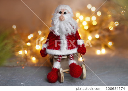 knitted toy Santa Claus sitting on wooden sites against a festive bokeh background knitted toy Santa Claus sitting on wooden sites against a festive bokeh background 108684574