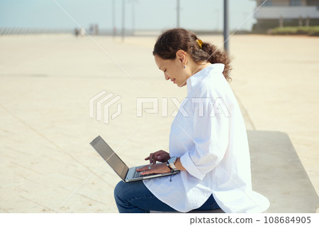 Side portrait of young business woman sitting outdoors with laptop on knees. Online messaging. Remote work. People. Job Side portrait of young business woman sitting outdoors with laptop on knees. Online messaging. Remote work. People. Job 108684905