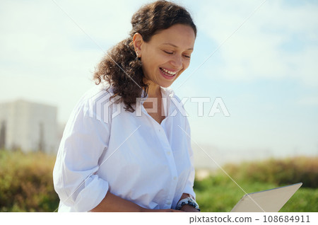 Beautiful multi ethnic young adult business woman smiling broadly while online working outdoors, using laptop Beautiful multi ethnic young adult business woman smiling broadly while online working outdoors, using laptop 108684911