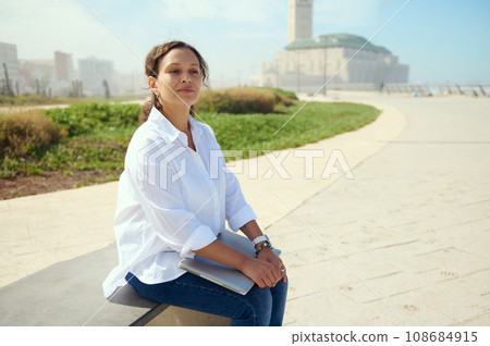 Confident business woman, freelance entrepreneur sitting on a bench outdoors, dreamily thinking on new startup project Confident business woman, freelance entrepreneur sitting on a bench outdoors, dreamily thinking on new startup project 108684915
