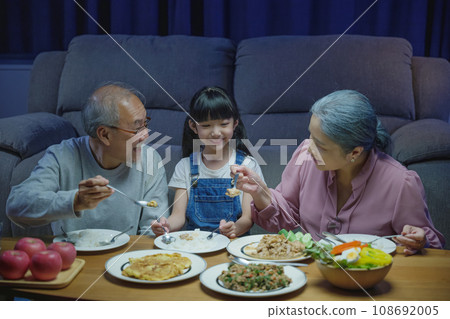 Happy Asian senior parent and child eating dinner food together in living room indoors, family grandmother grandfather and granddaughter dining on table and having fun during at home night time 108692005