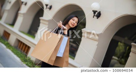 Happy beautiful young stylish woman asian with shopping bag while walking near mall on holiday Black Friday Happy beautiful young stylish woman asian with shopping bag while walking near mall on holiday Black Friday 108694034