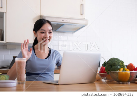 Happy woman asian in stylish tracksuit fresh smoothie shooting video with modern laptop in kitchen 108694132