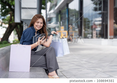 Woman using smartphone holding Black Friday shopping bag while standing on the stairs with the mall background Woman using smartphone holding Black Friday shopping bag while standing on the stairs with the mall background 108694381