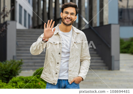 Indian man smiling friendly at camera, waving hands hello, hi, greeting or goodbye in city street 108694481