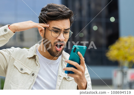 Surprised Indian man use smartphone loses, bad news, fortune loss, fail outdoors in city street Surprised Indian man use smartphone loses, bad news, fortune loss, fail outdoors in city street 108694519