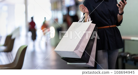 Woman using smartphone holding Black Friday shopping bag while standing on the stairs with the mall background 108694945