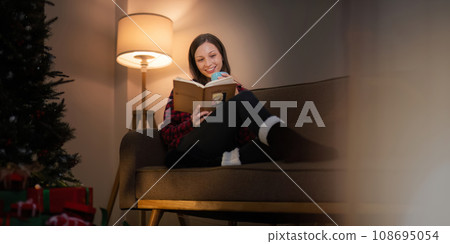 Smiling woman with a cup of tea and a book relaxing in side of a decorated Christmas tree in living room. concept holidays Christmas festive celebrate and tree Xmas seasonal 108695054