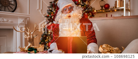 Surprised Santa Claus with glowing gift near christmas tree. New year and Merry Christmas concept Surprised Santa Claus with glowing gift near christmas tree. New year and Merry Christmas concept 108698125
