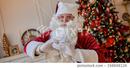 Santa Claus forming shape of heart with fingers near fireplace and christmas tree with gifts 108698126