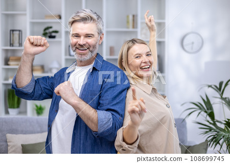 Happy senior couple dancing in their cozy living room, smiling looking at camera, happy long marriage, husband and wife spending weekend at home. 108699745