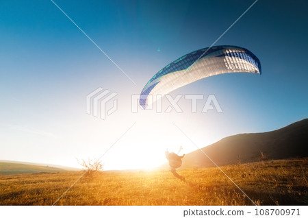 Launching a speedwing from a mountain. A paraglider is preparing to take off from a mountain, running along the yellow autumn grass against the sun Launching a speedwing from a mountain. A paraglider is preparing to take off from a mountain, running along the yellow autumn grass against the sun 108700971