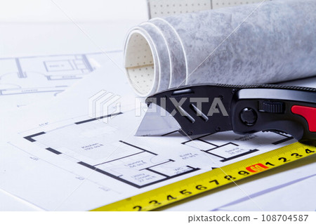 Laying flooring. Linoleum, a knife and a house plan on the builder's table. Taking measurements for laying the floor. Laying flooring. Linoleum, a knife and a house plan on the builder's table. Taking measurements for laying the floor. 108704587